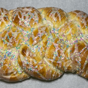 May include: A braided loaf of golden-brown bread, glazed with white icing and sprinkled with colorful nonpareils. The baked good has a glossy appearance and is likely a sweet bread or pastry.