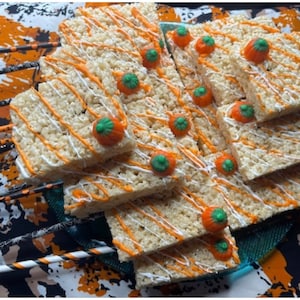 May include: A stack of square rice crispy treats, decorated for Halloween. Each treat is drizzled with orange and white icing and topped with a candy pumpkin. The treats are arranged on a teal plate with Halloween-themed straws.