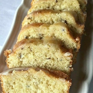 May include: Sliced loaf cake with a light yellow crumb and a white glaze. The cake is on a white, oblong serving dish. The cake slices have visible dark specks, possibly from ingredients like poppy seeds or herbs.