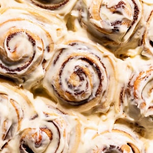 May include: Overhead shot of freshly baked cinnamon rolls in a metal baking pan. The rolls are swirled with a dark brown cinnamon filling and generously covered in a thick, creamy white frosting. The warm tones and textures create an inviting image.