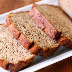 Puede incluir: Cuatro rebanadas de pan de plátano en un plato blanco. El pan es de color marrón claro y tiene una textura ligeramente desmenuzable.