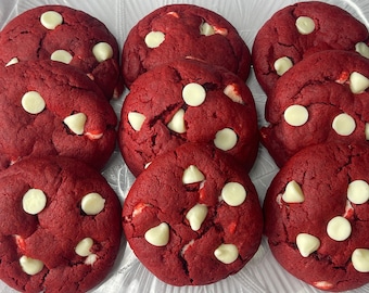 May include: Nine red velvet cookies with white chocolate chips are arranged in a clear container. The cookies are round and have a slightly cracked surface. The white chocolate chips are scattered throughout.