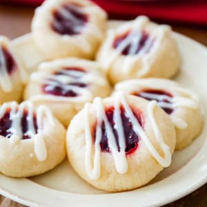 May include: A plate of thumbprint cookies with a dollop of red jam in the center and drizzled with white icing. The cookies are arranged on a cream-colored plate, with a few more cookies visible in the background.