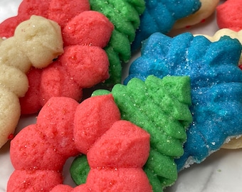 May include: Close-up of assorted colorful cookies. The cookies are shaped like Christmas trees and bows, and are decorated with red, green, blue, and white frosting and sprinkles. The cookies are arranged closely together.