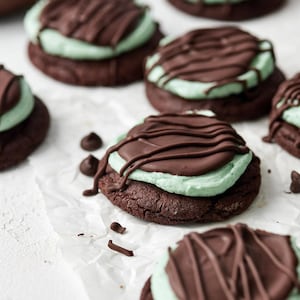 May include: A close-up of chocolate mint cookies with a chocolate drizzle on top. The cookies are arranged on a white parchment paper background.