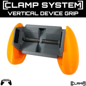 Op de afbeelding: Een verticale apparaatgreep met de tekst "CLAMP SYSTEM" en "VERTICAL DEVICE GRIP" in het wit. De greep is grijs met oranje handgrepen en een zwarte klittenbandriem. Het apparaat heeft een veermechanisme.