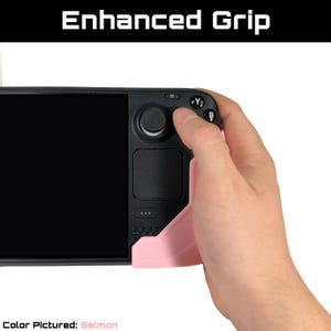 May include: A salmon-colored grip accessory for a handheld gaming device. The text "Enhanced Grip" is displayed at the top. The device is black with a screen and buttons. The grip is designed to improve handling.