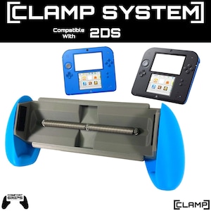 Könnte beinhalten: Ein graues Kunststoff-Klemmsystem mit hellblauen ergonomischen Seitengriffen und einer zentralen Metallspannfeder. Der Text lautet CLAMP SYSTEM, Compatible With 2DS, COMFORT GRIPS und CLAMP. Zwei 2DS-Handheld-Konsolen in Blau und Schwarz sind abgebildet.