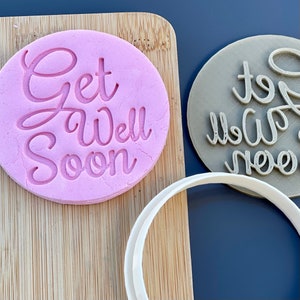 Peut inclure: Deux emportes-pièces en forme de biscuit, l'un rose et l'autre brun, avec le texte "Get Well Soon" en écriture cursive. Les emportes-pièces sont sur une surface en bois.