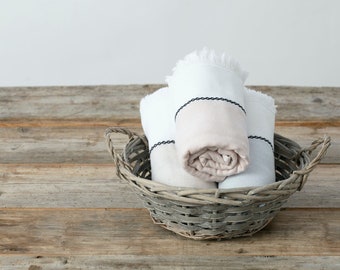 May include: Three rolled towels in a grey wicker basket. The towels are white with black trim and one has a light pink stripe.