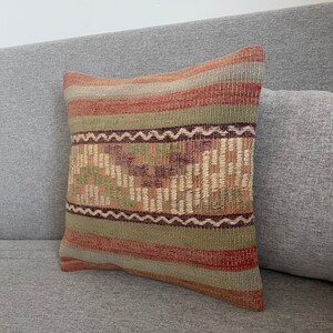 Op de afbeelding: Een decoratief kussen met een geweven patroon in tinten rood, groen, bruin en beige. Het kussen is gemaakt van een zachte, textuurrijke stof en heeft een rechthoekige vorm.