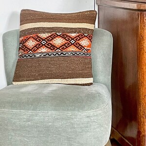 Op de afbeelding: Een bruin en beige kussen met oranje en zwarte geometrische patronen ligt op een lichtgroene fauteuil.