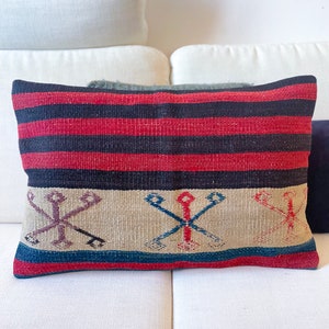 Peut inclure: Un coussin de décoration rayé rouge, noir et beige avec un motif géométrique. Le coussin est posé sur un canapé blanc.