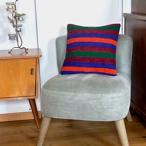 Op de afbeelding: Een grijze fauteuil met houten poten en een gestreept kussen in de kleuren rood, oranje, blauw en groen.