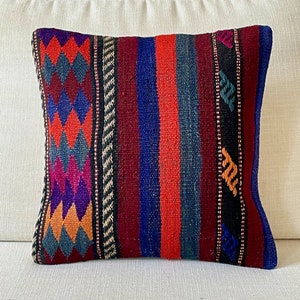Op de afbeelding: Een kleurrijk gestreept en gepatroneerd kussen met een donkerblauw, rood, oranje en paars kleurenschema. Het kussen is gemaakt van geweven stof en heeft een franje rand.