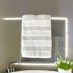 Puede incluir: Una toalla a rayas blancas y grises colgada de una barra cromada. La toalla está hecha de un material suave y absorbente. El entorno del baño incluye un grifo cromado y un jarrón blanco con vegetación.
