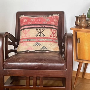 Op de afbeelding: Een bruine leren fauteuil met een gepatroneerd kussen. Het kussen heeft een geometrisch patroon in roze, beige en zwart. De fauteuil staat voor een houten bijzettafel met een koperen theepot en een kopje.