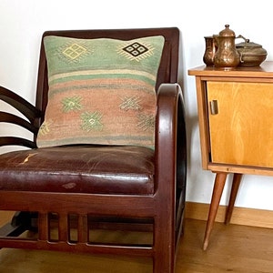 Op de afbeelding: Een bruine leren fauteuil met een gepatroneerd kussen. Het kussen heeft een groen, roze en wit geometrisch ontwerp. De stoel staat voor een houten bijzettafel met een lade en twee koperen kannen.
