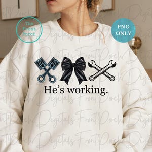May include: White sweatshirt with a black bow, two crossed wrenches, and two crossed pistons. The text "He's working." is printed below the design.