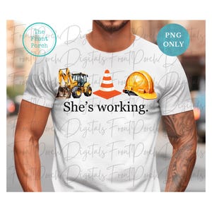 May include: A white t-shirt with a yellow excavator, an orange traffic cone, and a yellow hard hat. The text "She's working." is printed on the shirt.