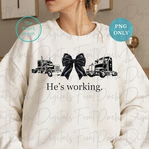 May include: A white sweatshirt with a black bow and two black semi-trucks on the front. The text "He's working." is printed below the trucks.