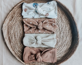 May include: Four fabric headbands in a woven basket. The headbands are tied in a knot and feature different patterns, including floral, polka dot, and solid colours. The headbands are in shades of brown, white, and beige.