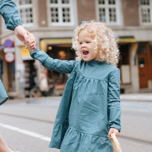 May include: A young girl with curly blonde hair wearing a teal blue dress with white polka dots and holding a straw bag. She is walking on a sidewalk and smiling.