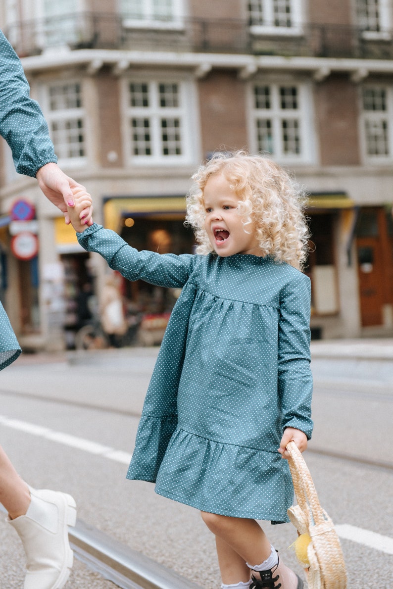 May include: A young girl with curly blonde hair wearing a teal blue dress with white polka dots and holding a straw bag. She is walking on a sidewalk and smiling.