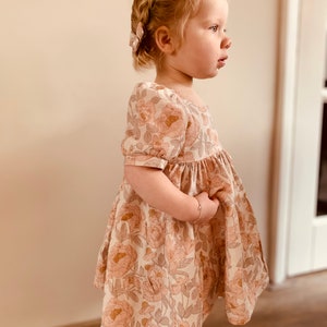 May include: A young girl wearing a white dress with a floral print. The dress has puffed sleeves and a gathered skirt. The girl has blonde hair in a braid and is wearing a hair clip.