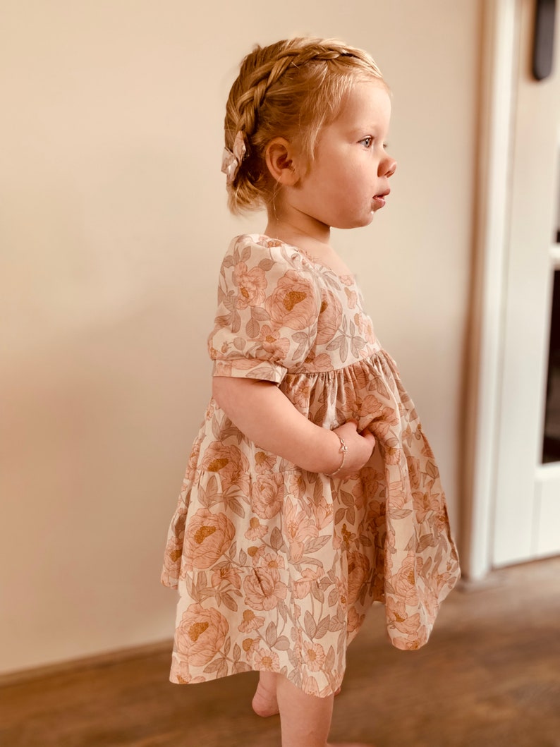 May include: A young girl wearing a white dress with a floral print. The dress has puffed sleeves and a gathered skirt. The girl has blonde hair in a braid and is wearing a hair clip.