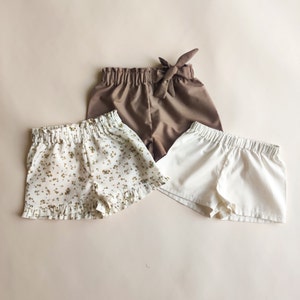 May include: Three pairs of shorts in different styles and colors. One pair is brown with a bow tie, one is white, and one is cream with a floral print.