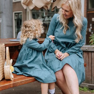 May include: A mother and daughter wearing matching teal blue dresses with white polka dots. The girl is wearing pink boots and the woman is wearing white ankle boots. They are sitting on a wooden bench outside.