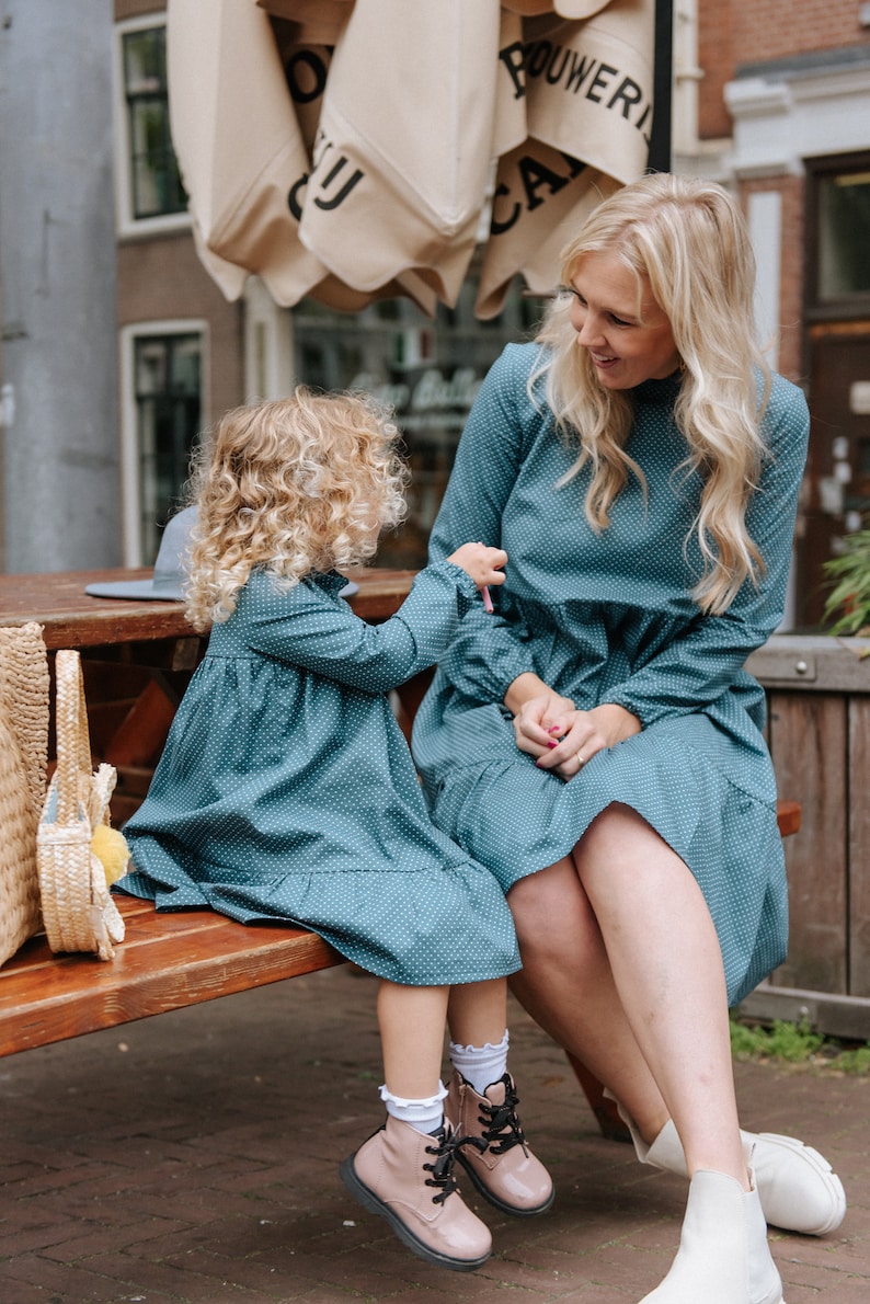 May include: A mother and daughter wearing matching teal blue dresses with white polka dots. The girl is wearing pink boots and the woman is wearing white ankle boots. They are sitting on a wooden bench outside.