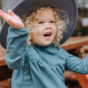 May include: A young girl with curly blonde hair wearing a teal blue dress with white polka dots and a gray wide-brimmed hat. She is smiling and looking up.