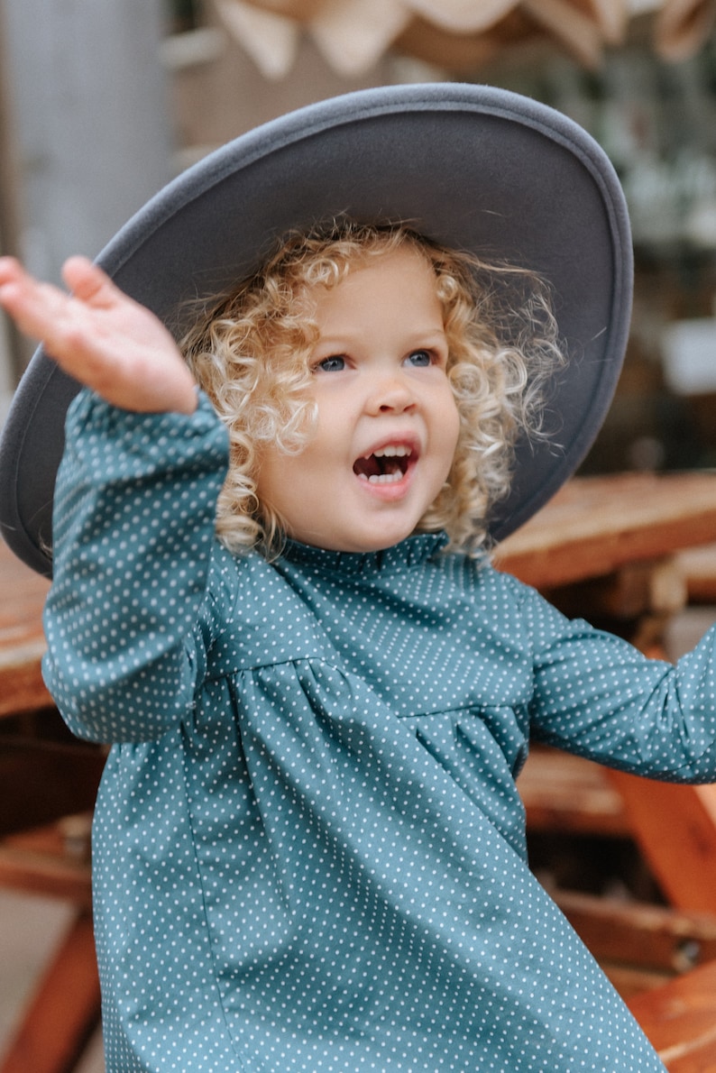 May include: A young girl with curly blonde hair wearing a teal blue dress with white polka dots and a gray wide-brimmed hat. She is smiling and looking up.