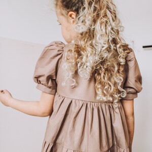 May include: A young girl wearing a brown tiered dress with puffed sleeves. Her hair is pulled back in a ponytail with a light brown scrunchie.