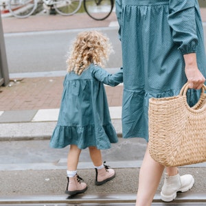 May include: A woman and a young girl wearing matching blue dresses with white polka dots. The woman is carrying a woven straw tote bag. They are walking on a sidewalk.