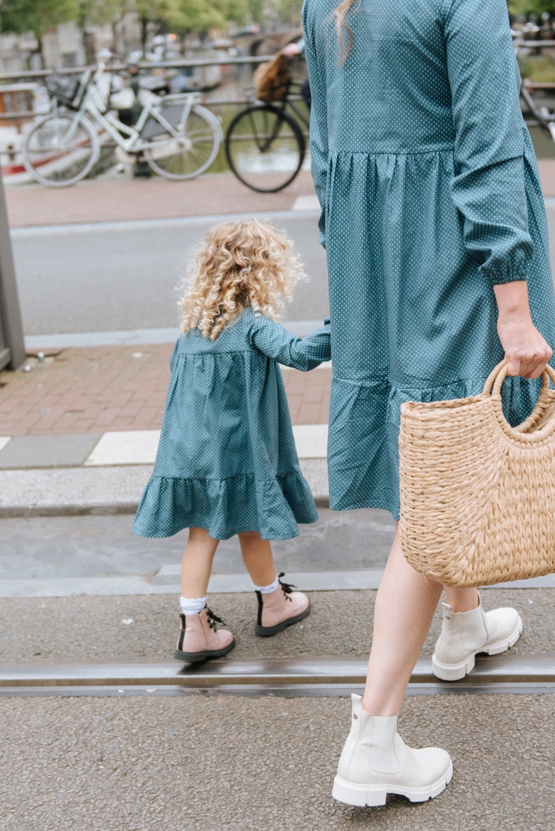 May include: A woman and a young girl wearing matching blue dresses with white polka dots. The woman is carrying a woven straw tote bag. They are walking on a sidewalk.