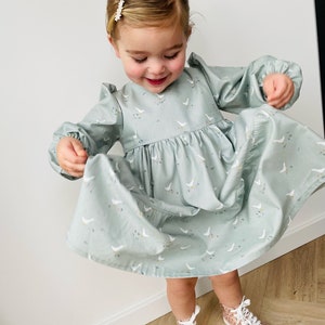 May include: A young girl wearing a light blue dress with a white goose print. The dress has long sleeves and a gathered skirt. She is wearing pink and white high-top sneakers.