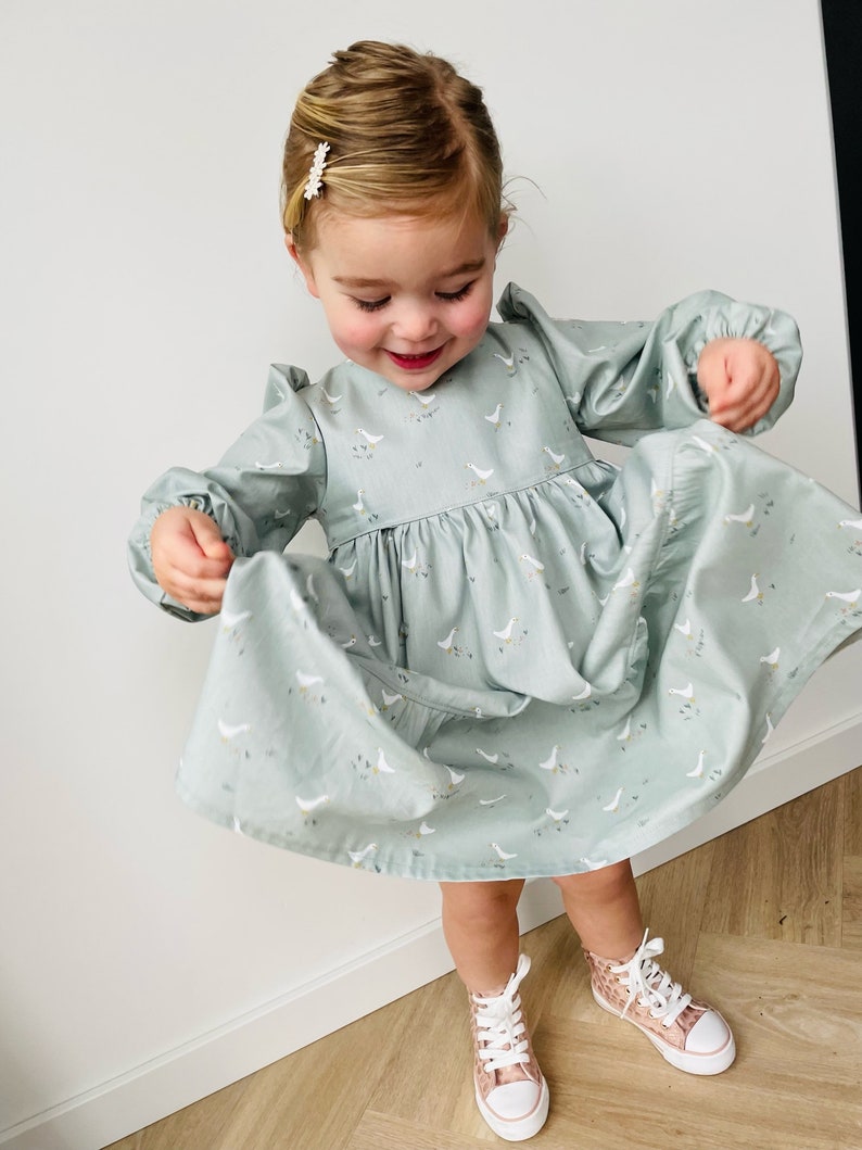 May include: A young girl wearing a light blue dress with a white goose print. The dress has long sleeves and a gathered skirt. She is wearing pink and white high-top sneakers.