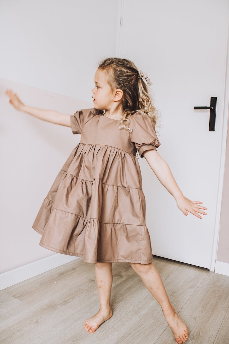 May include: A young girl wearing a brown tiered dress with puffed sleeves, standing on a wooden floor and facing away from the camera. She is wearing her hair in a braid with a small brown bow.