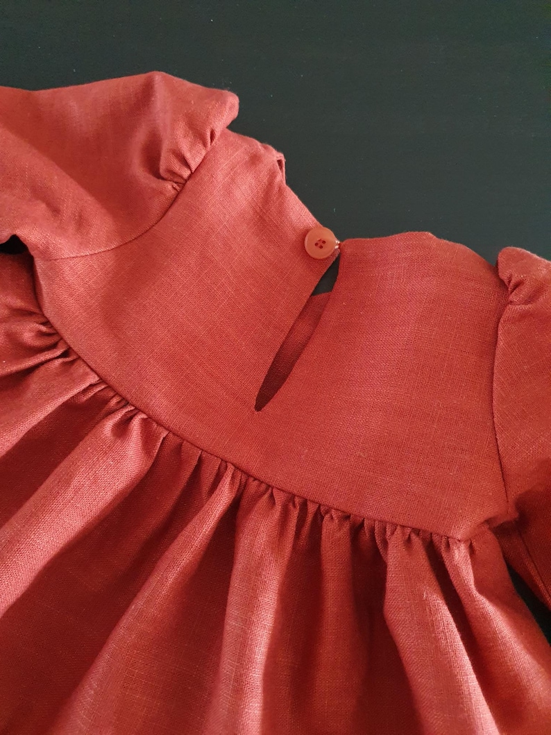 May include: A rust-colored linen dress with a button closure at the back of the neck. The dress has long sleeves and a gathered skirt.