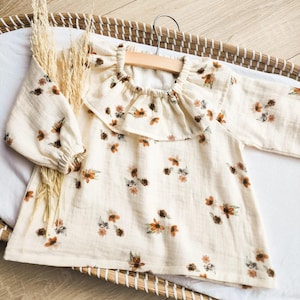 May include: A white baby blouse with a frilled collar and a floral print in shades of brown and orange.