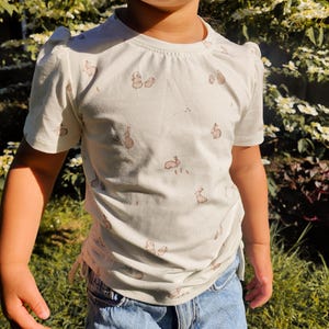 Pode incluir: Uma camiseta branca com um padrão de pequenos coelhos marrons. A camisa de manga curta tem um detalhe lateral franzido e é combinada com shorts jeans. A camisa tem mangas bufantes.