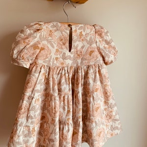 May include: A tiered floral dress with a keyhole back and puff sleeves. The dress is made of a light-weight fabric with a floral print in shades of pink, brown, and white.