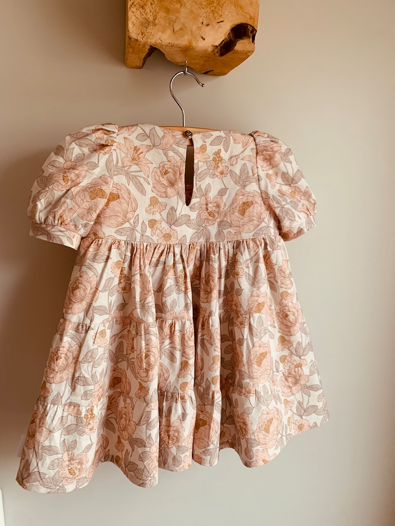 May include: A tiered floral dress with a keyhole back and puff sleeves. The dress is made of a light-weight fabric with a floral print in shades of pink, brown, and white.