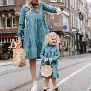 May include: A woman and a young girl wearing matching blue dresses with white polka dots. The woman is wearing a light blue hat and carrying a woven straw tote bag. The girl is holding a small round woven straw bag. They are standing on a street in front of a brick building.