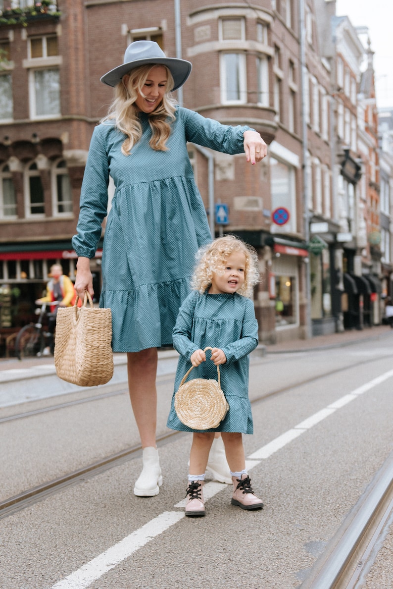 May include: A woman and a young girl wearing matching blue dresses with white polka dots. The woman is wearing a light blue hat and carrying a woven straw tote bag. The girl is holding a small round woven straw bag. They are standing on a street in front of a brick building.