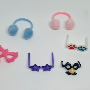 May include: A set of six colorful plastic toy accessories for dolls. The set includes a pair of pink earmuffs, a pair of blue earmuffs, a pair of purple star-shaped glasses, a pair of white glasses with red and blue paw prints, a pink masquerade mask, and a black mask with a mustache and a nose.