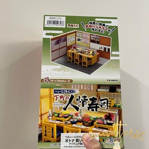 May include: A box for a Japanese sushi restaurant miniature set. The box is yellow and features a picture of the miniature set, which includes a sushi bar, tables, chairs, and sushi dishes. The text on the box reads "人情寿司" which translates to "Humanity Sushi".